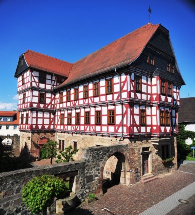 Hochzeitshaus. Blick aus dem Nachbarhaus der anderen Straßenseite. Zu sehen ist ein Blick auf den Hof hinter der Mauer. Die Mauer ist von einem kleinen Brunnen durchbrochen, so das von der Hof- als auch von der Straßenseite an den Brunnen herangetreten werden kann. Dann das Hochzeitshaus mit dem unteren Stockwerk aus Stein, die weiteren Stockwerke aus Fachwerk, weiß verputzte Gefache mit rot farbenen Holzbalken. Dach mit Tonziegeln gedeckt.