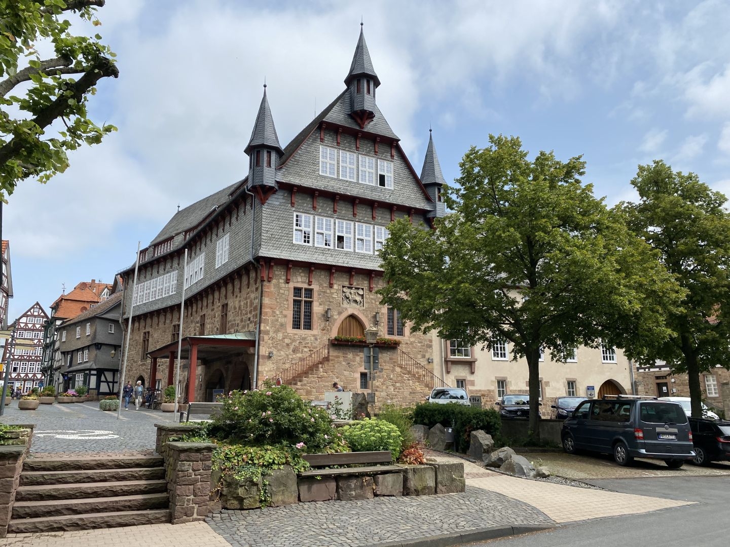 Rathaus mit Türmchen. Das untere Stockwerk ist aus Sandstein mit leicht rötlicher Farbe gebaut. Die beiden oberen Stockwerke sind aus Fachwerk mit blaugrauem Schiefer verkleidet und von einer Reihe von Fenstern versehen, hinter denen sich die Büros der Verwaltung befinden. Das Dach ist ebenfalls mit blaugrauem Schiefer gedeckt. Im rechten Bereich sind vor dem Gebäude noch zwei Bäume zu sehen.