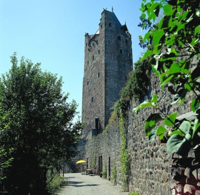 Grauer Turm. Blick entlang der Stadtmauer mit grauem Basaltstein, Mittig ragt der Graue Turm hervor. Links noch ein Strauch aus einem Garten der in die Straße wächst. Zu sehen ist noch der Treppenaufgang für den Besuch im Turm.