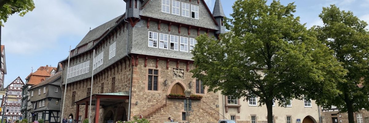 Rathaus mit Türmchen. Das untere Stockwerk ist aus Sandstein mit leicht rötlicher Farbe gebaut. Die beiden oberen Stockwerke sind aus Fachwerk mit blaugrauem Schiefer verkleidet und von einer Reihe von Fenstern versehen, hinter denen sich die Büros der Verwaltung befinden. Das Dach ist ebenfalls mit blaugrauem Schiefer gedeckt. Im rechten Bereich sind vor dem Gebäude noch zwei Bäume zu sehen.